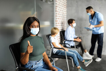Afro-american young woman in protective face mask showing thumb up, sitting at clinic, waiting for vaccination against coronavirus. Multiracial patients different ages getting immunization