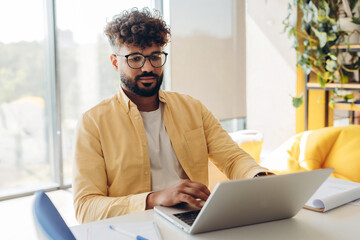 Handsome, confident Middle Eastern man wearing eyeglasses, using laptop sitting in modern office