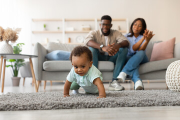 A happy baby boy crawls on the soft rug as he learns to walk in the living room. His proud parents sit on the sofa, clapping and encouraging their little one during this special moment.