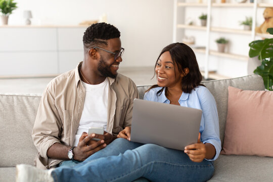 A joyful black couple relaxes on their couch at home, using a laptop and smartphone. They engage in conversation while watching movies and enjoying each other's company during the weekend.