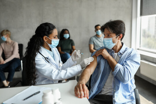 Male patient getting intramuscular shot against COVID-19 at clinic or vaccination center, middle-eastern woman doctor making injection in shoulder for caucasian man, wearing protective face masks - Powered by Adobe