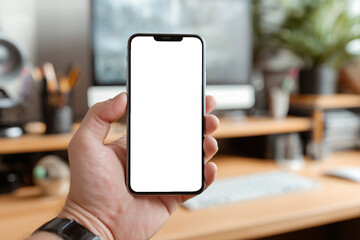 A close-up of a hand holding a smartphone with a blank black screen inside a modern home office workspace.