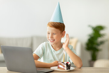 Cheerful redhead boy in a party hat waves at his laptop during an online birthday celebration. A birthday cake sits on his desk, capturing the moment of joy amid the coronavirus pandemic.