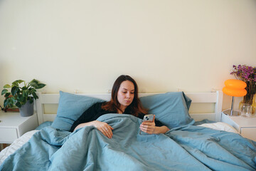 Obraz premium A young woman lies in bed in a cozy bedroom and looks at her phone. The girl checks her email and social media after waking up