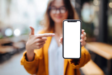 A young woman wearing glasses and a yellow jacket holds a smartphone with a blank black screen toward the camera while pointing at it in an indoor setting.