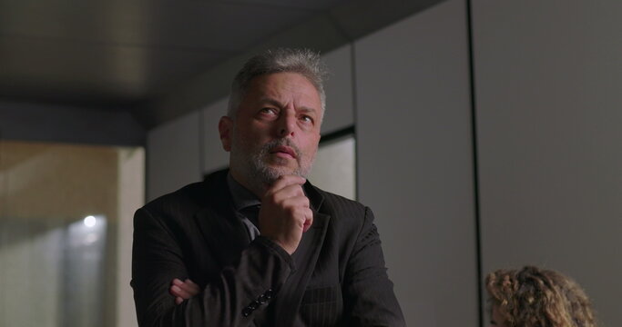 Close up of mature businessman with hand on chin and focused expression while considering decision during corporate meeting in modern office - Powered by Adobe