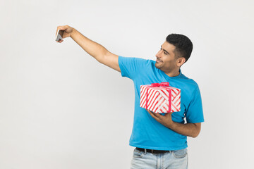 Positive man blogger holding mobile phone and present box, making selfie or having video call.