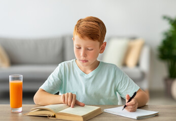 A concentrated redhead boy is sitting at his desk, reading a book and taking notes. He enjoys fresh juice full of vitamins while doing his homework in a bright room with a cozy atmosphere.
