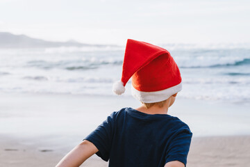 Сhild wearing red Santa hat stands on beach and looks toward the ocean waves. Christmas spirit and coastal scenery. Back view of boy in red hat on the ocean shore.
