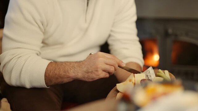 A man is sitting in front of a fireplace, cutting a piece of cheese with a knife. Concept of warmth and comfort, as the man enjoys a simple snack in a cozy setting