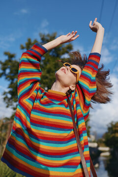 Woman in a rainbow sweater and yellow sunglasses outdoors, joyful and stretching arms toward blue sky; casual outfit, mid-twenties model enjoying fashion lifestyle on a sunny day.