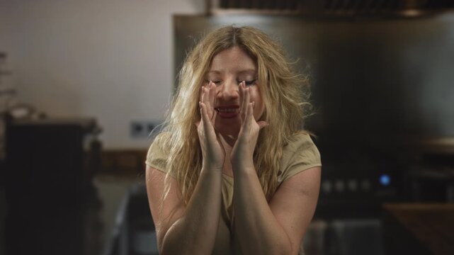 Young blonde woman cupping hands to mouth shouting in a building kitchen with messy hair and visible forearms; playful announcement.