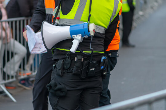 A security officer with a megaphone supervises a street event among barriers and spectators
