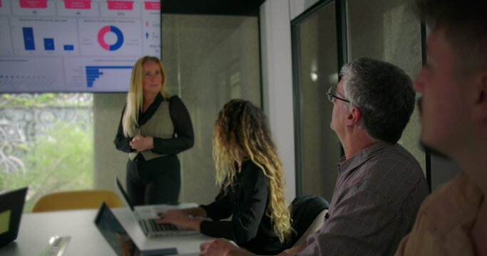 Businesswoman standing in front of screen while explaining data points to engaged team seated around table in modern office setting