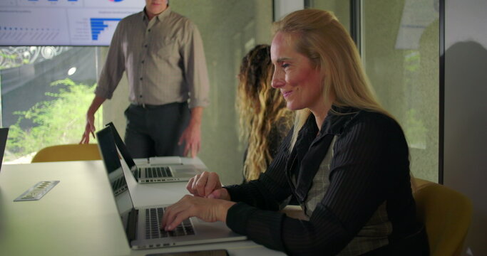 Businesswoman typing with slight smile during meeting as coworkers collaborate beside her in glass walled corporate conference room - Powered by Adobe