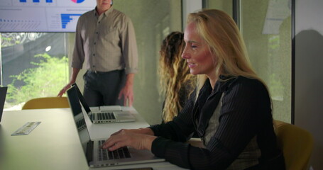 Businesswoman typing with slight smile during meeting as coworkers collaborate beside her in glass walled corporate conference room