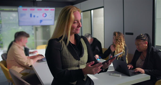 Businesswoman using digital tablet while team collaborates in background during office meeting, modern workplace with technology-driven productivity and professional focus