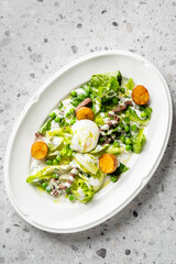 A fresh, gourmet Caesar salad with poached egg, roasted potatoes, green peas, and meat on a white platter. Healthy eating, fine dining closeup