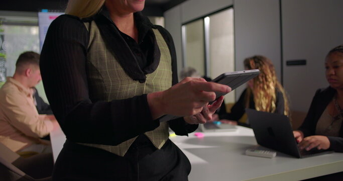 Close-up of businesswoman using tablet during office meeting, collaborative workspace with team members working in background, highlighting technology and leadership focus