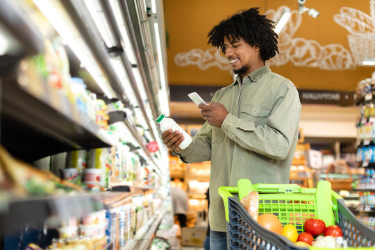 A man stands in a grocery store aisle, holding a milk carton while checking prices on his phone. A shopping cart filled with groceries sits beside him.