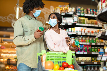 A couple is shopping in a grocery store, both wearing face masks. They are looking at products in their cart and enjoying a light conversation. Shelves are stocked with various items.