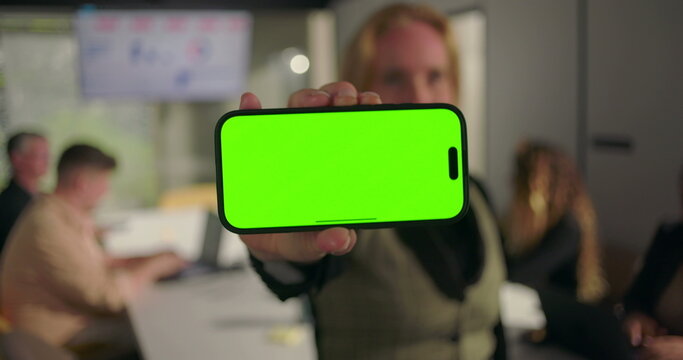 Close-up of hand holding smartphone with green screen, blurred business team working in modern conference room with charts on screen