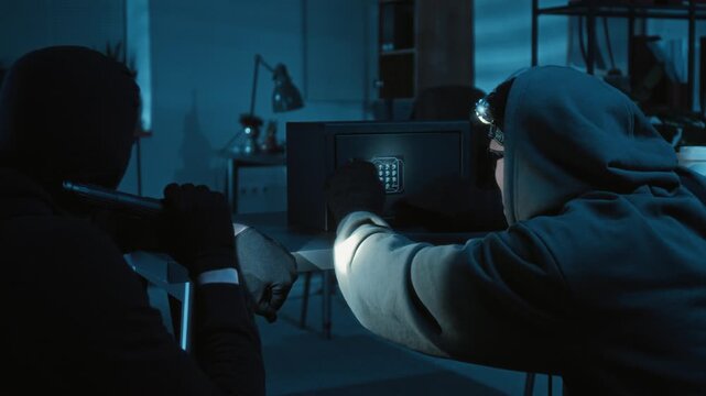 Young criminal with headlamp cracking electronic safe and taking stack of dollar bills from it with mouth opened in surprise while his accomplice shining torch around dark office