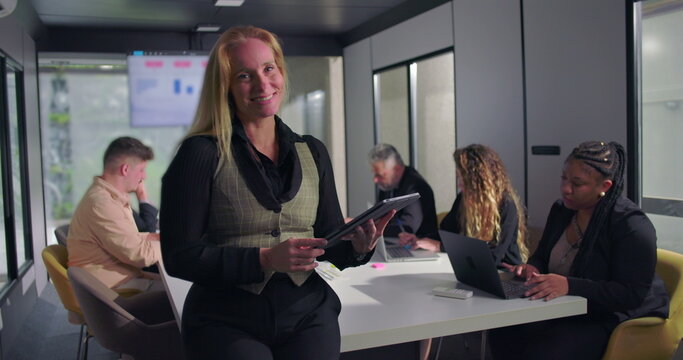 Cheerful female executive holding tablet while standing at head of conference table during office meeting, team working in background in productive and positive environment