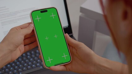 A woman is holding a smartphone featuring a green screen, set against a modern laptop on her desk. She appears focused and engaged in her work.