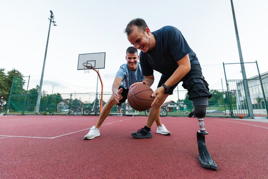 A man with a prosthetic leg dribbles a basketball while playing with a friend at an outdoor court during daylight. - Powered by Adobe