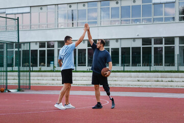 A man with a prosthetic leg enjoys a game of basketball on an outdoor court with a friend during sunny weather.