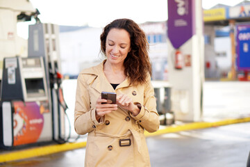 A middle-aged woman with curly hair and a trench coat smiles while using her cell phone with an out-of-focus gas station in the background. © Inspiraciones