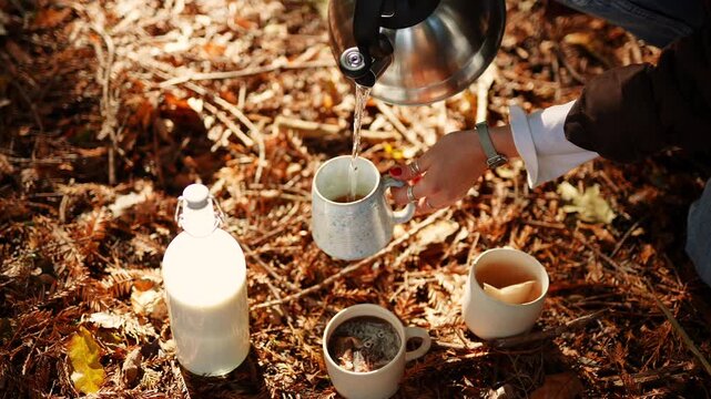 A person is pouring tea into a cup from a kettle. There are three cups and a bottle of milk on the ground