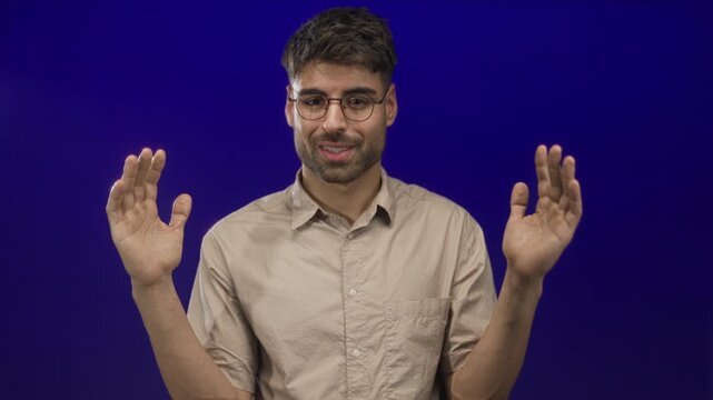 Young hispanic man wears glasses and holds palms apart in a blue studio wearing a beige shirt; clarity calm.