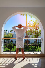 Obraz premium man in bathrobe stands on the terrace of a luxury house overlooking the sea and palm trees, rear view.