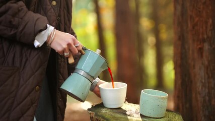A woman is pouring coffee into a white cup from a coffee pot. The scene is set in a forest, with trees in the background. The woman is wearing a brown coat and a white shirt - Powered by Adobe