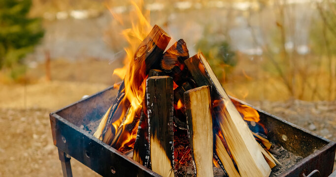 Glowing wood logs burning intensely inside traditional metal brazier, preparing for outdoor grilling with warm orange red flames - Powered by Adobe