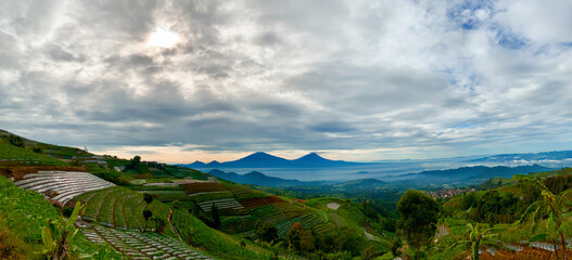 Panoramic view of lush green hills and terraced landscapes overlooking a serene lake with distant Merbabu and Merapi mountains under a cloudy sky