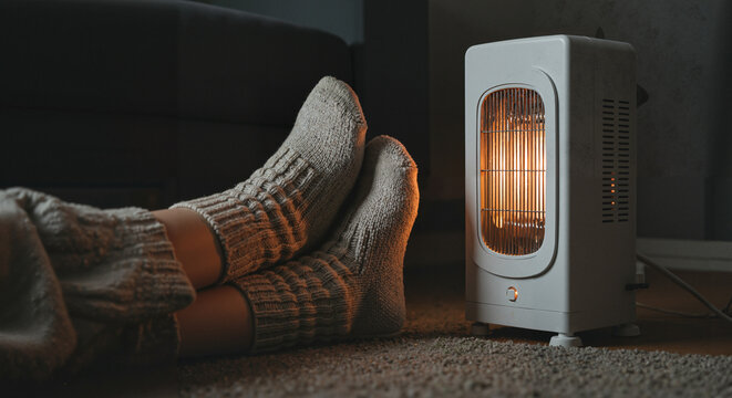 Cozy feet in warm socks near electric heater on carpeted floor  