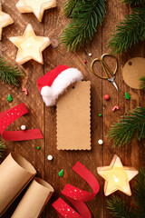 Blank gift tag with mini Santa hat on wooden table. Christmas flat lay with star candles, red ribbon, and fir branches. Copy space for holiday wishes.