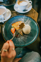 A person is eating a dessert with a fork. The dessert is served on a blue plate with a fork and a spoon. The dessert is topped with whipped cream and strawberries
