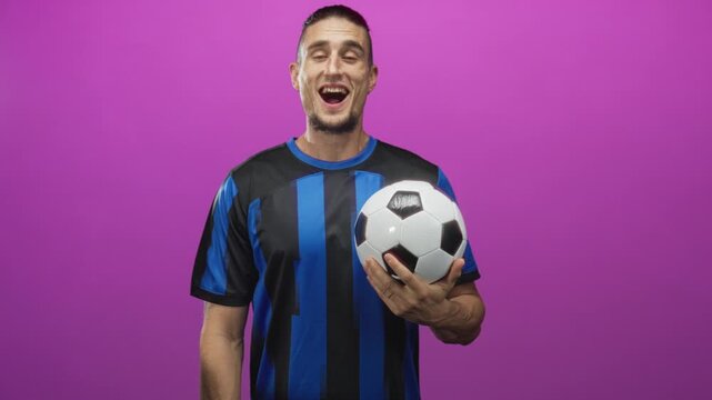 Man holding soccer ball with hand, smiling in studio wearing blue striped jersey; joyful celebration.