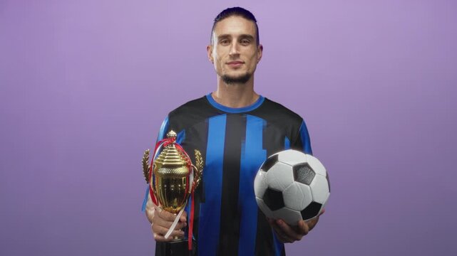 Man holds trophy and soccer ball in studio while wearing blue black jersey and smiling at camera; victory pride.