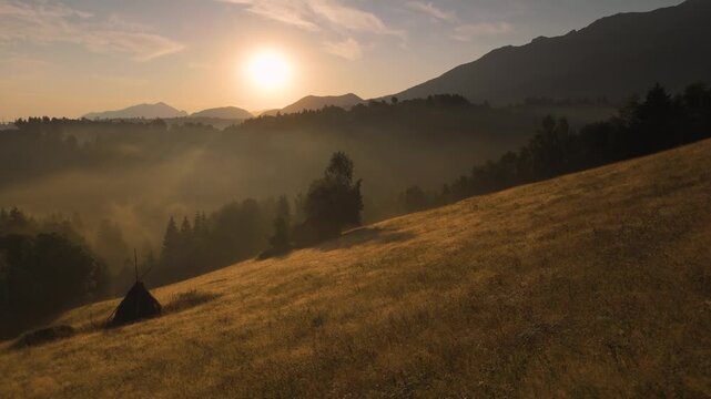 Transylvanian Carpathian Mountains sunrise over dry summer hillside meadow near Simon village, misty forest valley with moving fog and golden light in early August