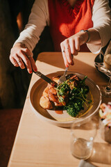 A woman is eating a meal with a knife and fork. The plate has meat and vegetables. The woman is wearing a white shirt and a red sweater