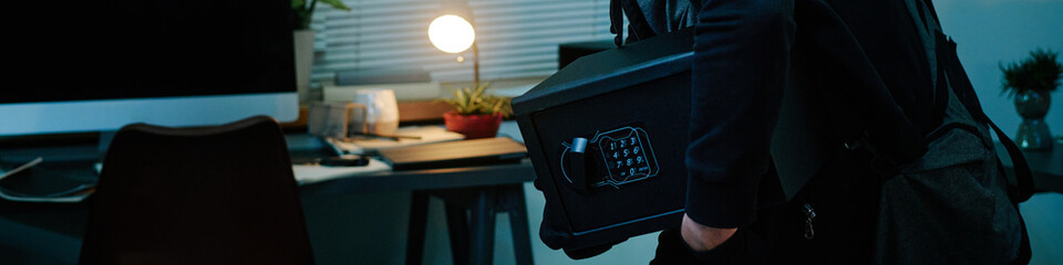 Website banner of middle aged man carrying safe while sneaking through office at night, holding...