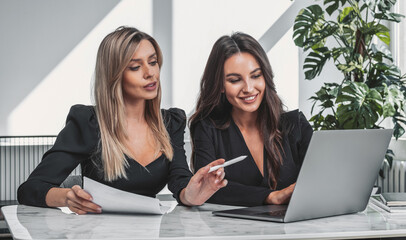 Two women in formal attire working together at a marble desk, using a laptop and paperwork, in a well-lit office with plants, concept of teamwork