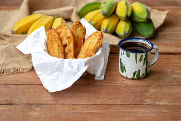 Fried bananas (Pisang Goreng) served with black coffee in a rustic enamel mug, set on a wooden table with fresh bananas on burlap in the background. Tropical snack/dessert.