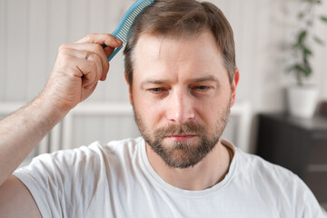 Obraz premium Handsome man combing brushing hair sitting at home light white bedroom. Guy styling grooming his hair with a comb. Self-care. Softly lit indoor space. Portrait. Male haircare morning routine concept