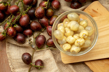 Top-down view of peeled Matoa (Pometia pinnata) flesh in a glass bowl on a wooden board, placed next to fresh purple fruit clusters on a rustic burlap background.